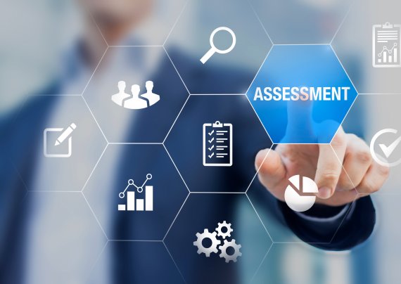 A person blurred out in the background with graphic content over the person. Icons are all around such as a magnify glass, charts, group of people. The focus if the design is pointing to a highlighted word of Assessment.