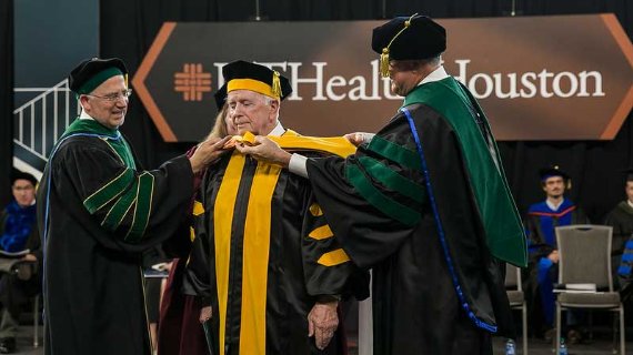 Dr. Colasurdo assisting in the robing of a graduate student on stage with the UTHealth Houston logo behind
