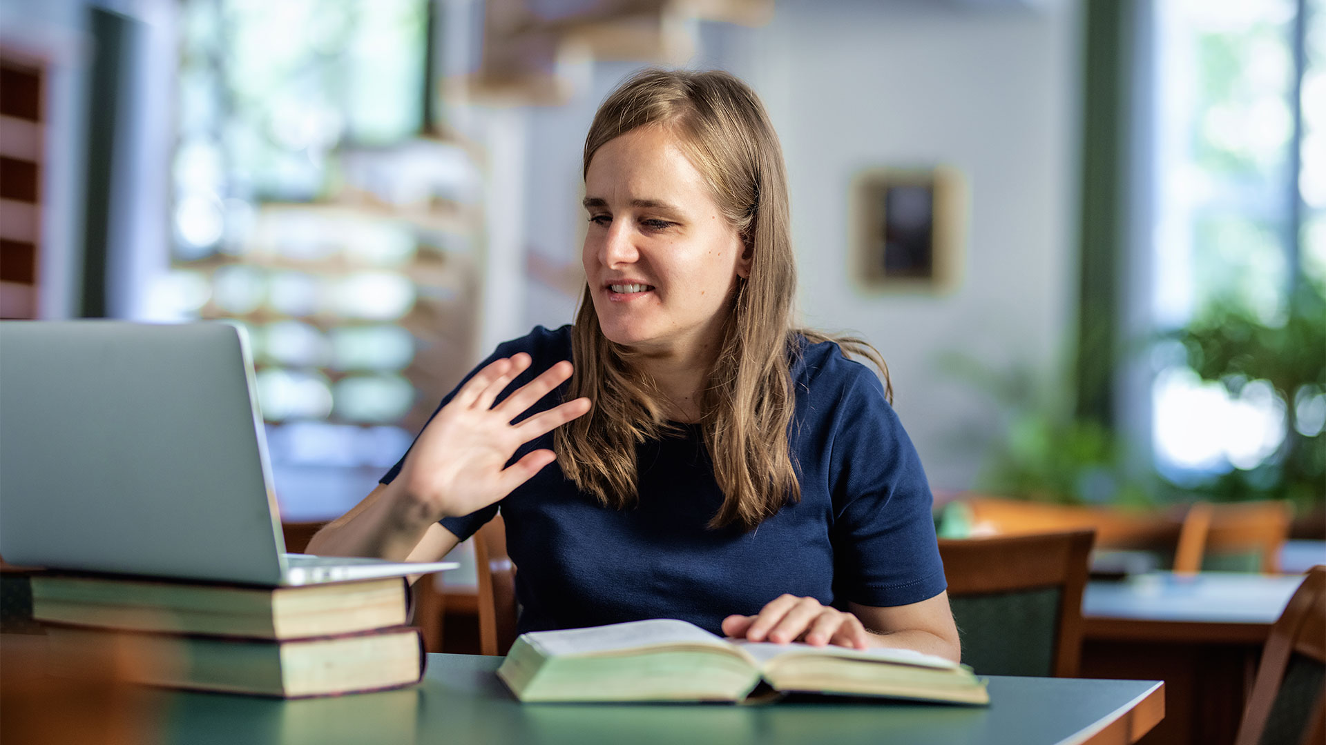 Woman waving during a video call on a laptop, with books and an open braille book on the table