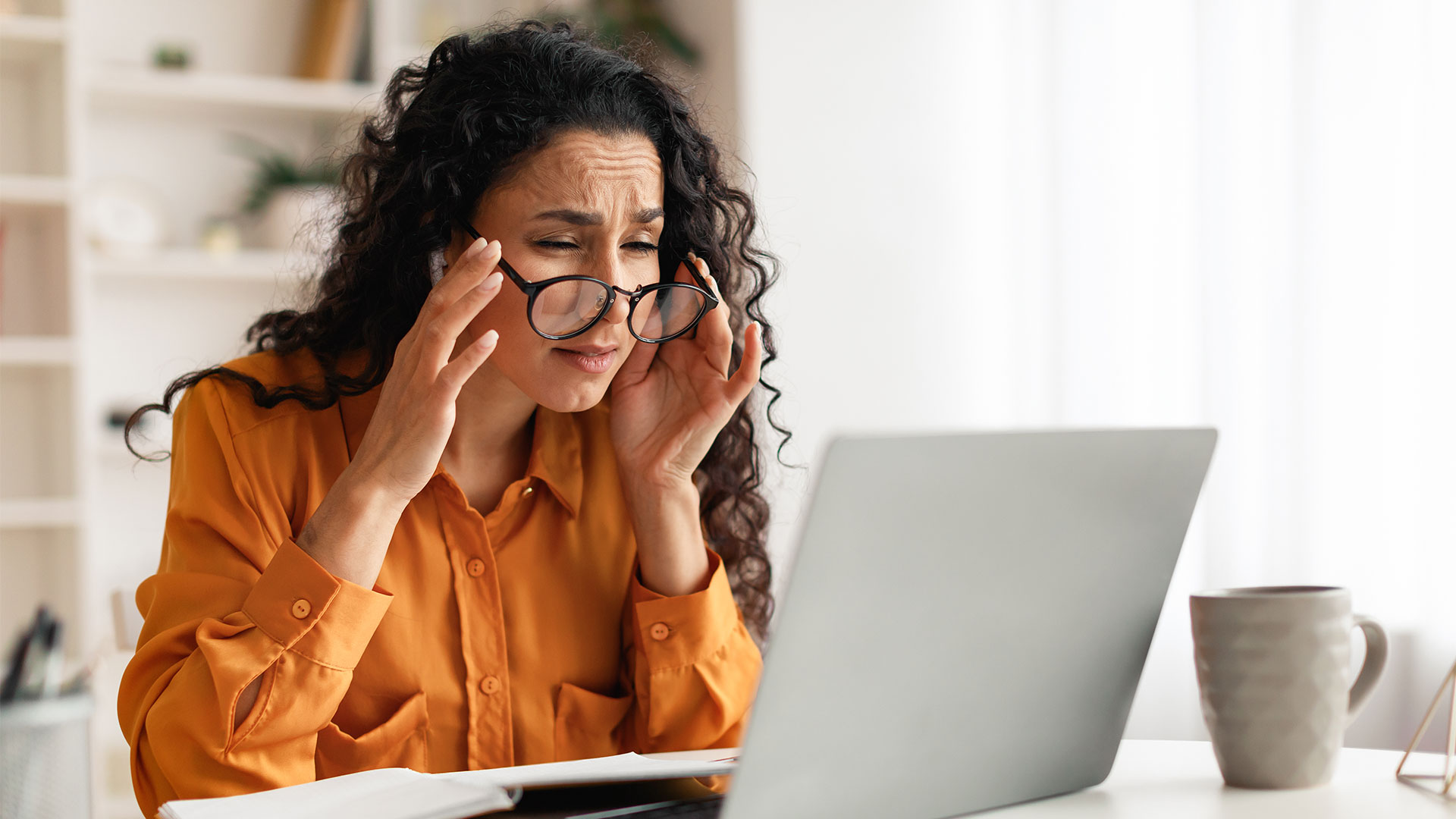 woman with glasses on, removing them below her eyes while squinting at a laptop screen
