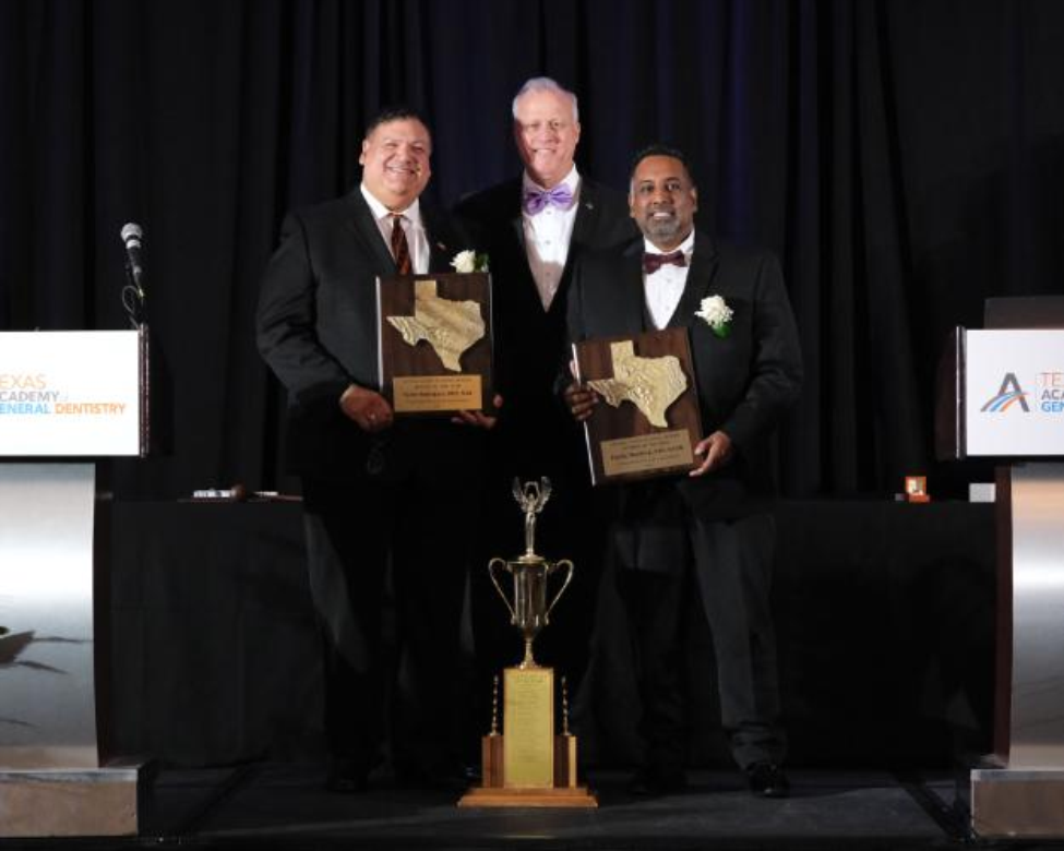 Three men stand on stage at the Texas Academy Awards. Two hold Texas-shaped plaques as 2025 Texas Dentist of the Year™ co-recipients on either side of the award presenter. A large trophy rests at their feet.