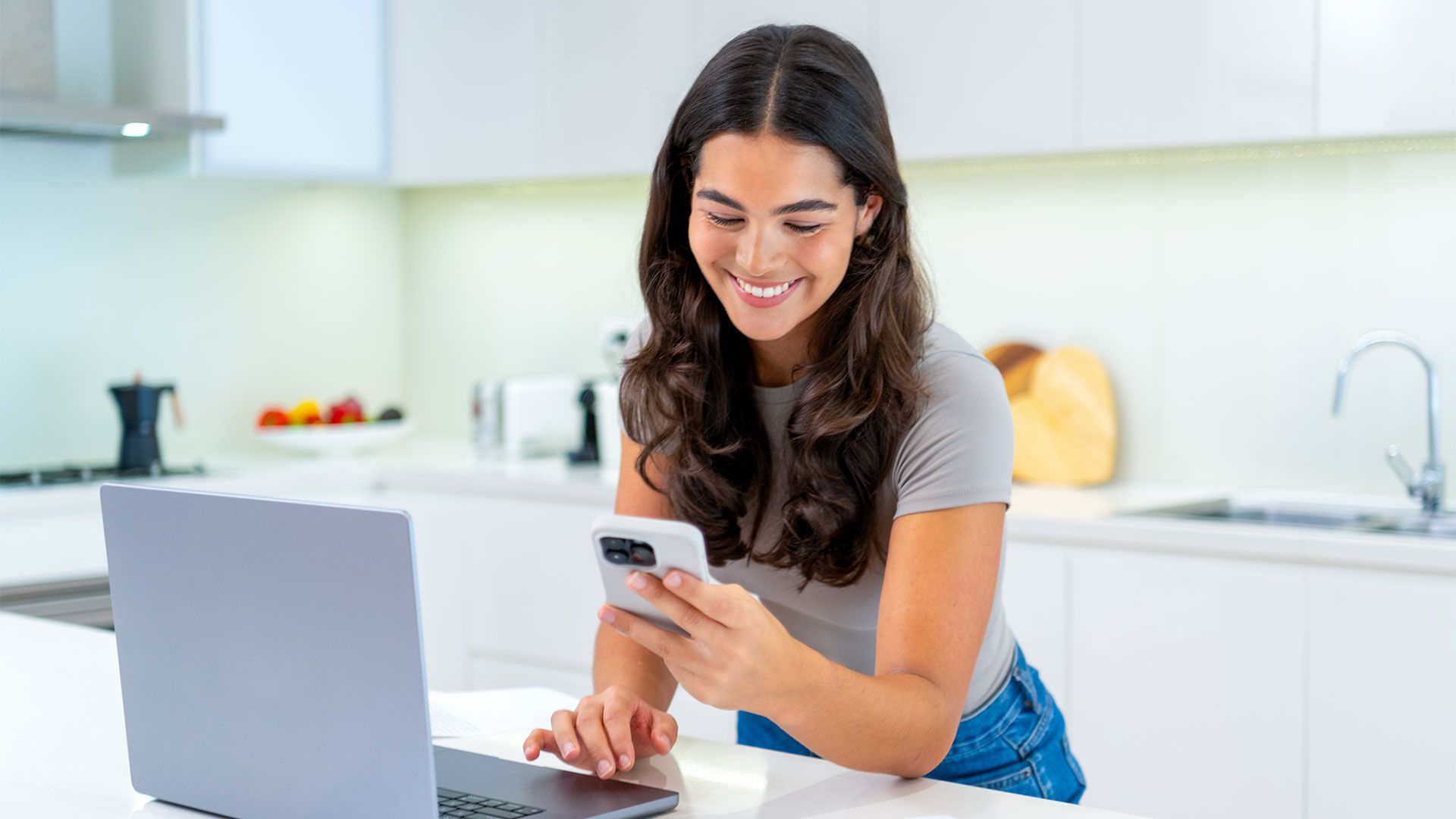 Woman holding her phone and smiling in the kitchen with a laptop in front of her