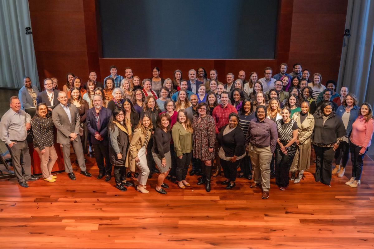 Large group photo shot from above with everyone in casual dress standing on a stage with hardwood floor.