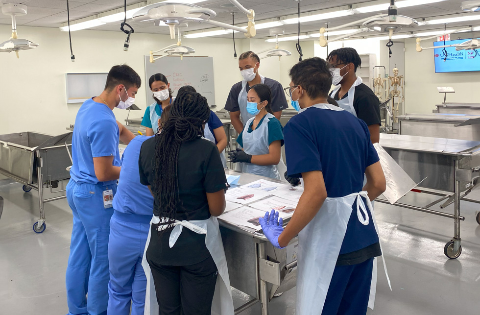 Group of individuals in scrubs and masks gathered around a table reviewing anatomical materials in a medical lab.