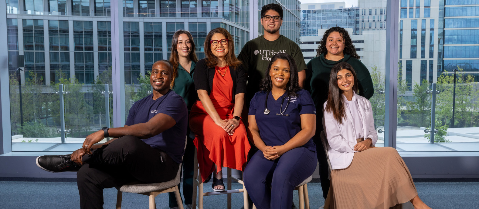 Group of eight UTHealth Houston Employees seated and standing indoors in front of large windows with city buildings in the background.